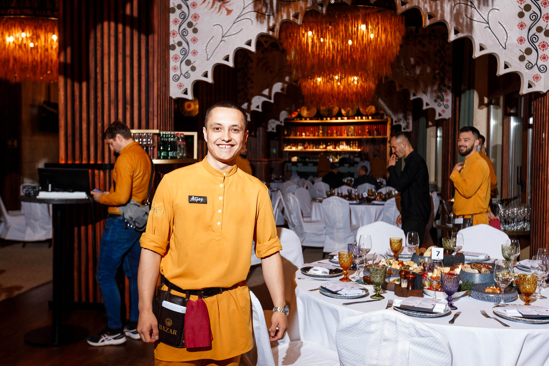 Официант в желтой униформе улыбается на камеру фотографу в ресторане the Bazar в Красной поляне