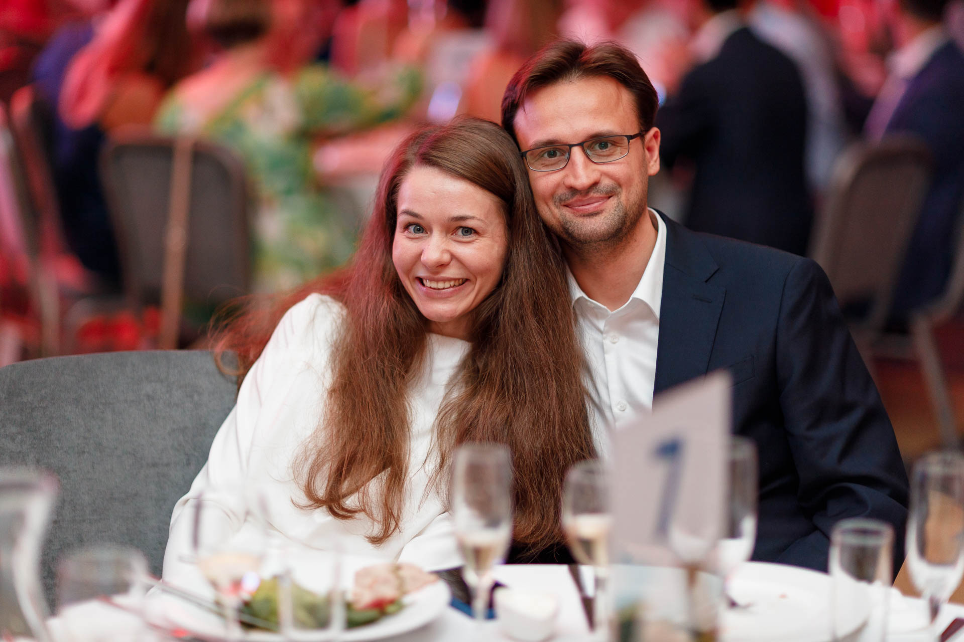 Пара, сидящая за столиком во время официального мероприятия и улыбающаяся в камеру
