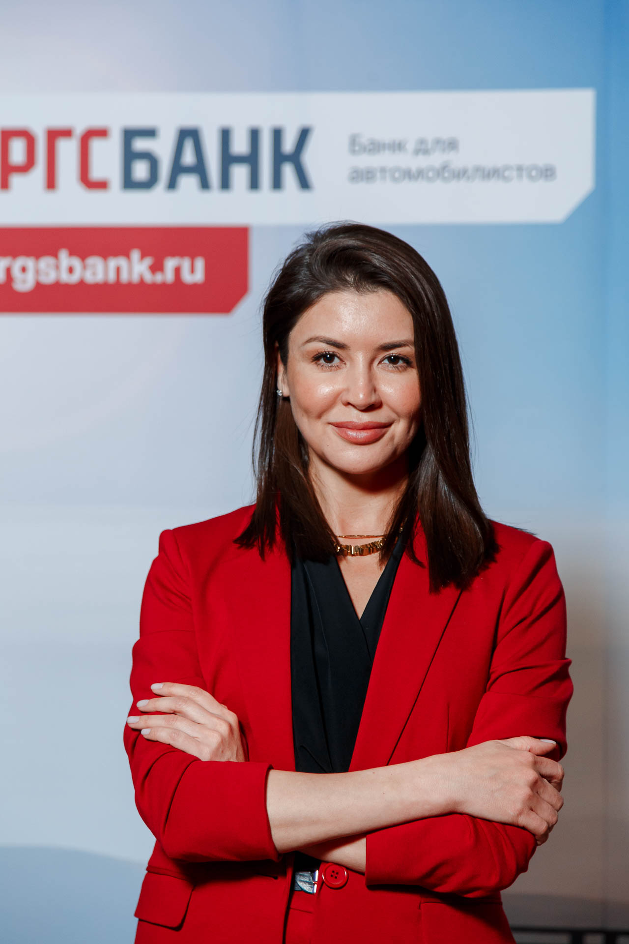 женщина в красном пиджаке, стоящая со скрещенными руками, улыбающаяся в камеру на рекламном фоне банка