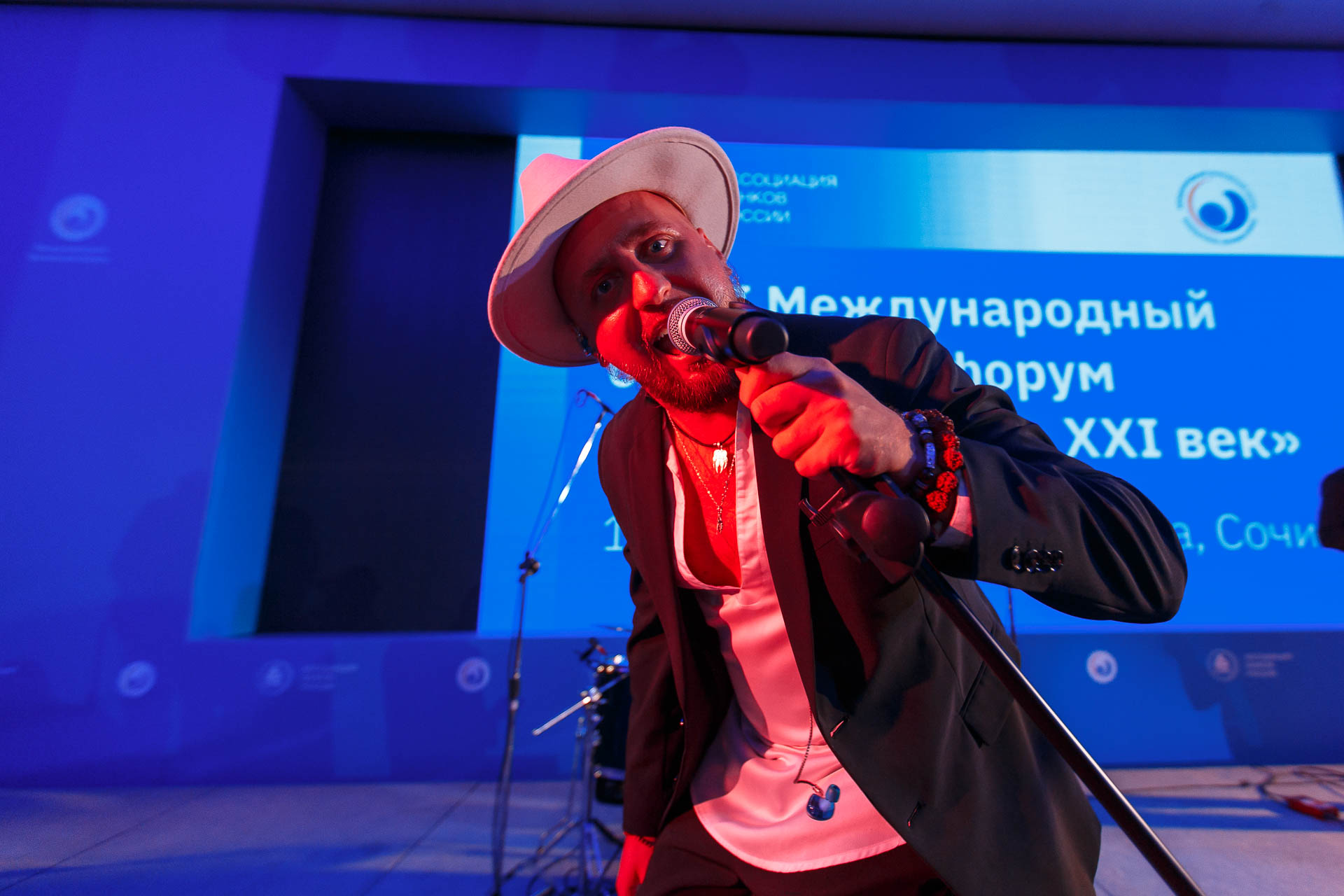 исполнитель, поющий в микрофон на сцене, освещенной ярким сценическим освещением, с экраном, отображающим детали конференции в Сочи