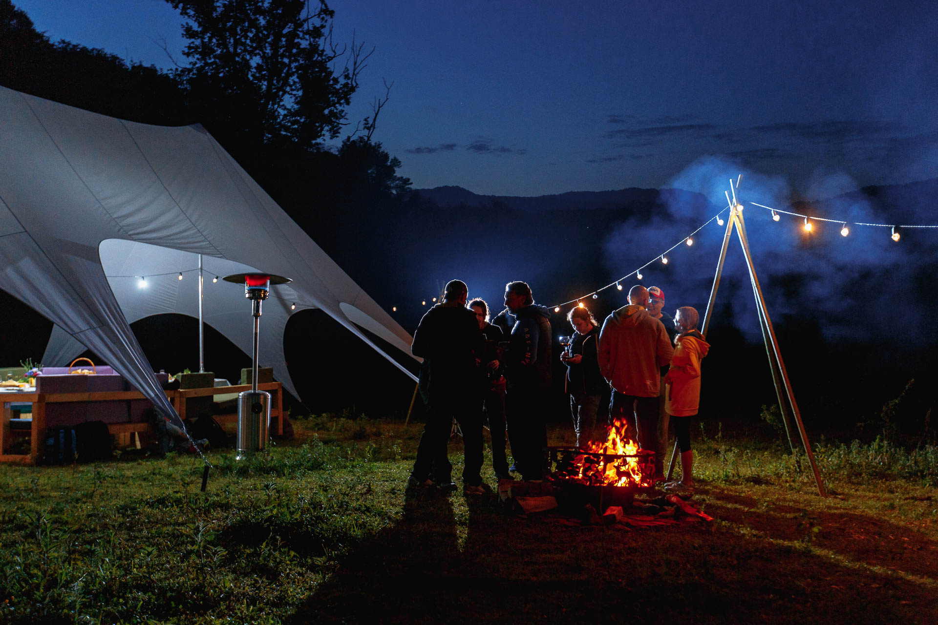 Группа друзей, наслаждающихся теплым костром спокойным вечером, с большой палаткой и гирляндами на улице