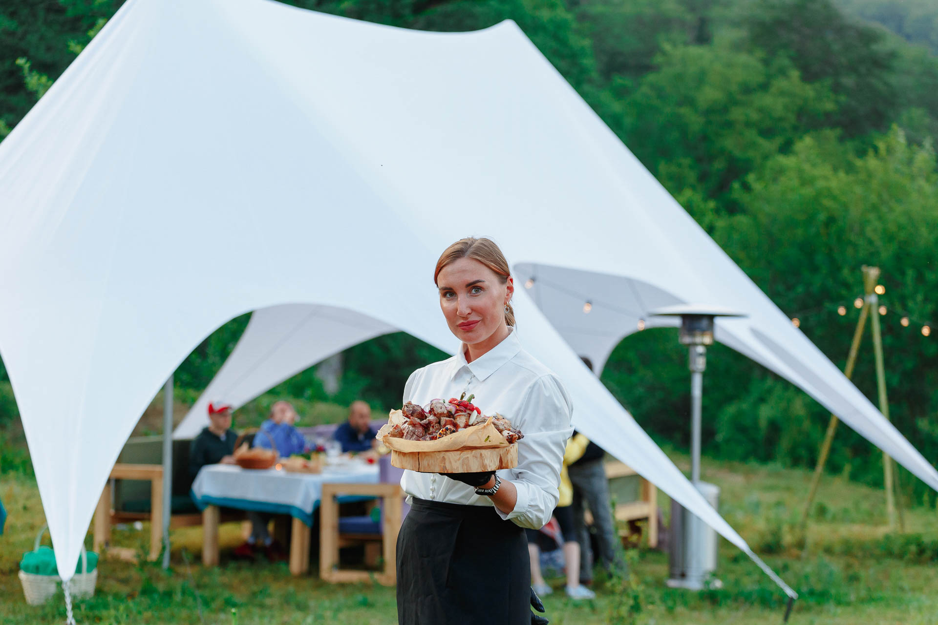 Официантка с подносом еды на мероприятии на открытом воздухе с белой палаткой и гостями на заднем плане